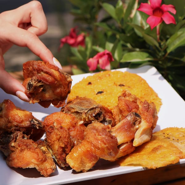 A hand picks up a piece of crispy fried chicken from a plate with yellow rice and plantains.